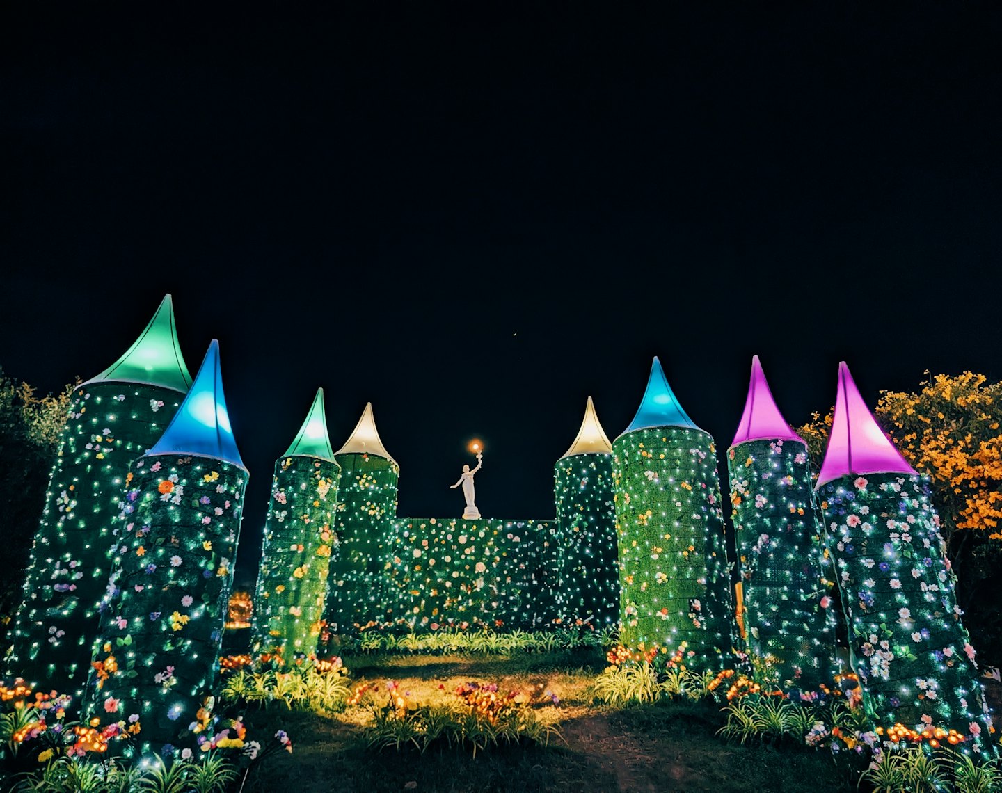 White statue holding a torch stands among illuminated floral towers.